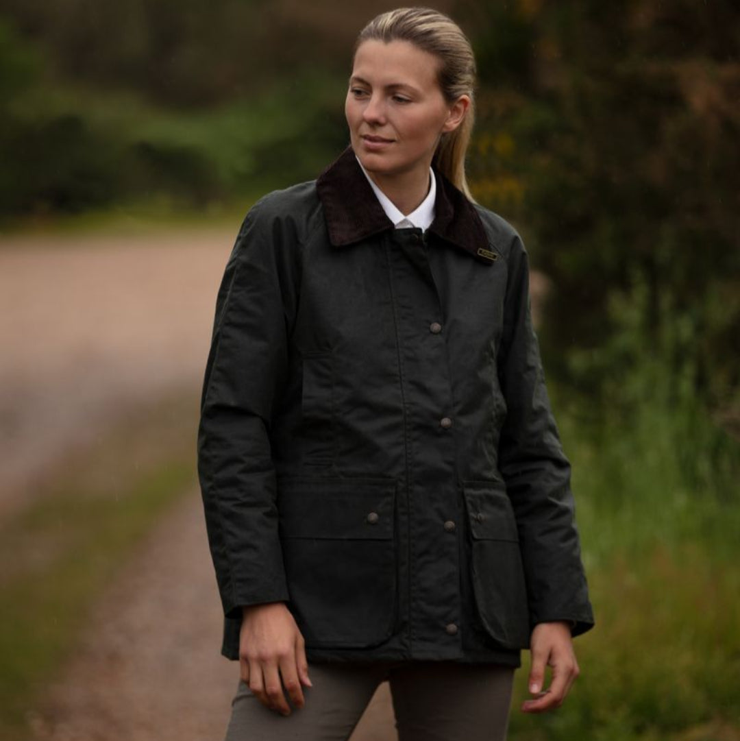 A woman wearing a dark green waxed cotton jacket with a corduroy collar, detachable hood, and elasticated cuffs.