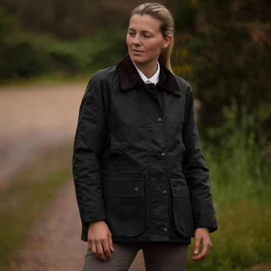 A woman wearing a dark green waxed cotton jacket with a corduroy collar, detachable hood, and elasticated cuffs.